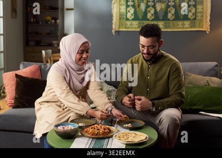 Junge Erwachsene muslimische Frau im Hijab und junger erwachsener Mann, der zusammen auf dem Sofa sitzt und zusammen Mahlzeiten teilt, lächelnd und genießt traditionelle Speisen im häuslichen Setting Stockfoto