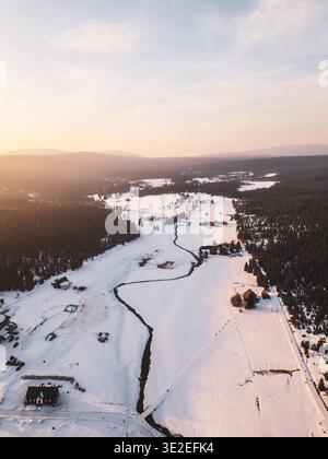 Schnee bedeckt das Dorf Jizerka, umgeben von Bäumen und Hügeln. Die Sonne untergeht, wirft Licht auf den sich windenden Fluss und die offenen Felder und schafft eine Winterszene in den Bergen. Stockfoto
