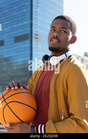 Afroamerikaner, der auf dem städtischen platz steht und orangefarbenen Basketball hält, Senfjacke trägt Stockfoto
