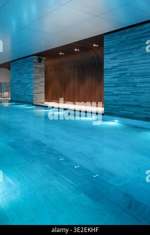 Leerer Innenpool mit Holzlamellenwand. Elegante Innenausstattung mit beleuchtetem Pool, strukturierten Steinsäulen und einer geschwungenen Holzwand. Stockfoto