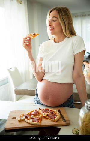 Glückliche schwangere Frau, die zu Hause Pizza isst. Schwangerschaftsbegehren, Mutterschaftsleben, Ernährung und pränatales Wellness-Konzept. Stockfoto
