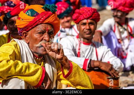 Ein älterer Mann in gelber Tunika und orangefarbenem Turban raucht ein Bidi, während er zusammen mit anderen Teilnehmern am Bundi Utsav Festival sitzt. Stockfoto
