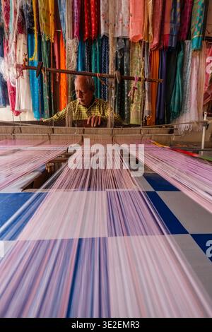 Ein einheimischer Weber in Bundi betreibt einen traditionellen Handwebstuhl und schafft farbenfrohe Textilien, umgeben von lebendigen hängenden Seidensaris. Stockfoto