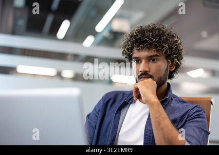 Nahaufnahme eines jungen Indianers, der nachdenklich an einem Schreibtisch sitzt und auf einen Laptop-Bildschirm blickt. Stockfoto