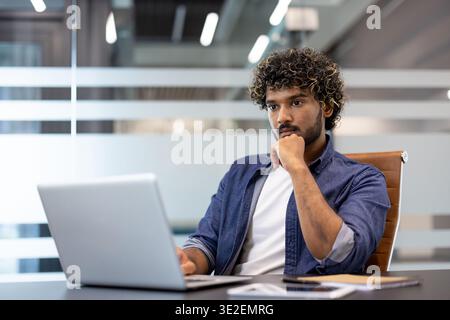 Ein nachdenklicher und konzentrierter junger Indianer sitzt an einem Schreibtisch und schaut ernsthaft auf seinen Laptop-Bildschirm. Stockfoto