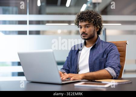 Ernsthafter und konzentrierter junger Indianer, der an einem Schreibtisch im Büro sitzt und an einem Laptop arbeitet. Stockfoto