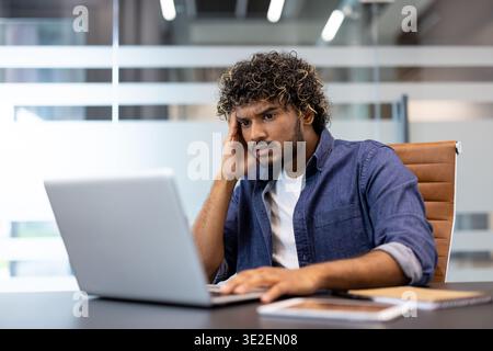 Ein verärgerter junger Indianer sitzt an einem Schreibtisch im Büro und schaut frustriert auf den Laptop-Bildschirm. Stockfoto