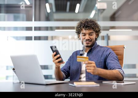 Ein lächelnder junger Indianer sitzt am Schreibtisch, benutzt Handy und Kreditkarte. Stockfoto