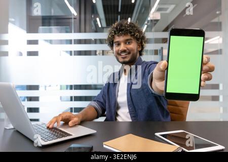 Ein lächelnder indischer junger Mann sitzt am Schreibtisch und zeigt einen grünen, leeren Bildschirm des Mobiltelefons vor die Kamera. Stockfoto
