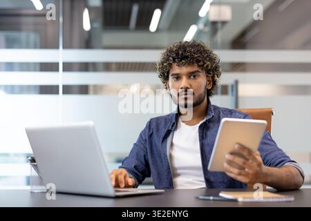 Porträt eines jungen ernsthaften Indianers, der im Büro mit einem Laptop sitzt. Er hält ein Tablet in der Hand und blickt auf die Kamera. Stockfoto