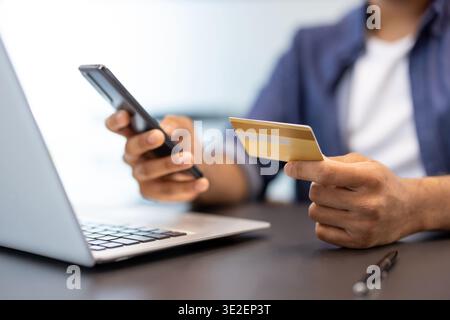 Ein Nahfoto der Hände eines jungen Mannes in einem Büro, der vor einem Laptop sitzt und eine Kreditkarte und ein Telefon hält. Stockfoto