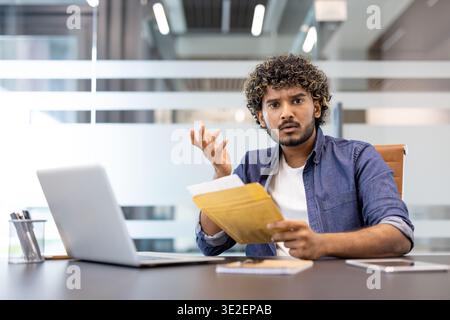 Ein verärgerter junger Indianer sitzt an einem Schreibtisch im Büro, hält einen offenen Brief mit einem Umschlag in der Hand und schaut enttäuscht in die Kamera. Stockfoto