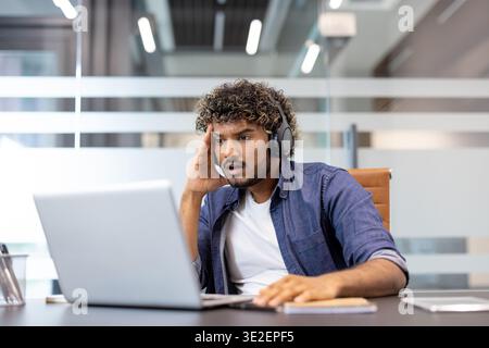Schockierter junger Indianer, der am Schreibtisch saß und Kopfhörer trug und überraschend auf den Laptop-Bildschirm schaute. Stockfoto