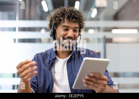 Muslimischer junger Mann, der im Büro mit der Kamera spricht, ein Headset trägt und ein Tablet benutzt. Stockfoto