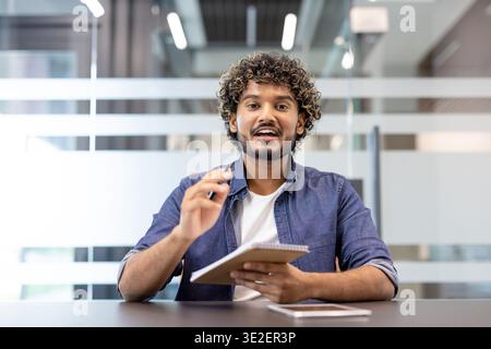 Junger indischer Lehrer, der vor der Kamera im Büro sitzt, ein Notizbuch hält, erklärt und mit der Kamera spricht. Stockfoto