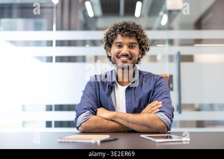 Porträt eines jungen indischen Mannes in einem Jeanshemd, der an einem Schreibtisch im Büro sitzt und in die Kamera lächelt. Stockfoto