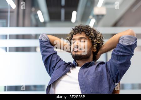Ein entspannter junger Indianer sitzt im Büro an einem Schreibtisch in einem Stuhl, Hände hinter dem Kopf, Augen geschlossen und ruhen. Stockfoto