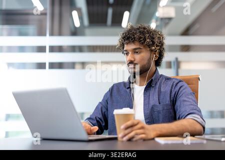 Ernsthafter und fokussierter junger Indianer, der im Büro mit Kopfhörern auf einem Laptop arbeitet, auf einem Stuhl sitzt und Kaffee trinkt. Stockfoto