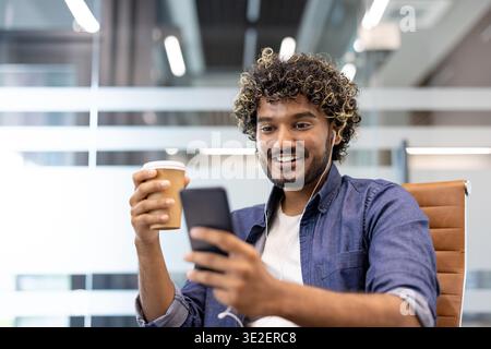 Ein lächelnder junger Indianer ist im Büro, trägt Kopfhörer, hält eine Tasse Kaffee und benutzt ein Handy. Stockfoto