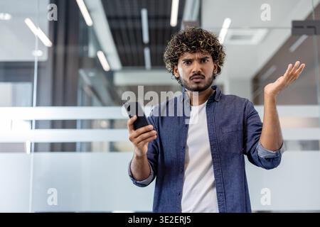 Porträt eines traurigen jungen Indianers in einem Büro, der ein Telefon hält und frustriert seine Arme spreizt. Stockfoto