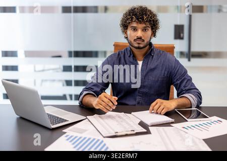 Porträt eines jungen ernsthaften Indianers, der an einem Schreibtisch im Büro sitzt und mit Dokumenten arbeitet und in die Kamera schaut. Stockfoto