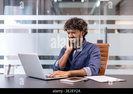 Ein junger Indianer, der vor Schmerzen grimmend ist, sitzt an einem Schreibtisch im Büro und hält seine Wange mit der Hand und spürt Zahnschmerzen. Stockfoto