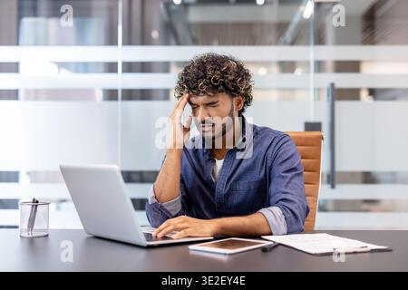 Müder junger Indianer sitzt an seinem Schreibtisch im Büro, verdreht vor Schmerz und Spannung, hält seinen Kopf mit der Hand. Stockfoto
