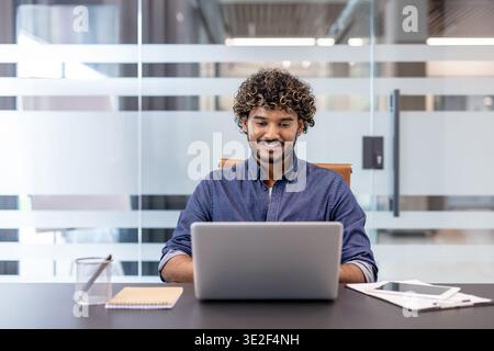 Ein lächelnder junger Indianer sitzt an einem Schreibtisch im Büro und arbeitet an einem Laptop. Stockfoto