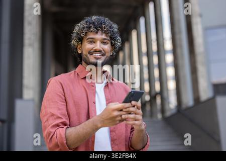 Porträt eines jungen Indianers in einem weißen Hemd, der draußen steht, ein Handy hält und in die Kamera lächelt. Stockfoto
