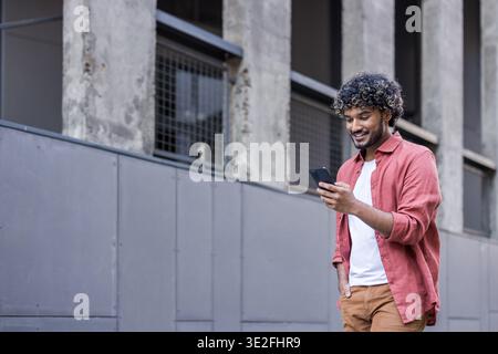 Junger indischer Mann in lässiger Kleidung, der auf einer Straße in der Nähe eines Gebäudes läuft und sein Handy benutzt. Stockfoto