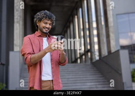 Ein lächelnder junger Indianer steht links neben dem Bürogebäude und benutzt das Handy. Stockfoto
