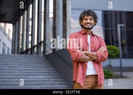 Porträt eines lächelnden jungen Indianers in einem roten Hemd, der selbstbewusst mit überkreuzten Armen auf der Brust steht und in die Kamera blickt. Stockfoto