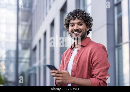 Nahaufnahme eines jungen muslimischen Mannes, der draußen steht, ein Telefon hält und in die Kamera lächelt. Stockfoto
