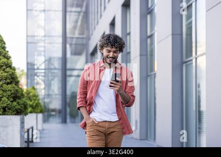 Ein junger, selbstbewusster muslimischer Mann läuft eine Straße entlang, benutzt sein Handy mit einem Lächeln und hält seine Hand in der Tasche. Stockfoto