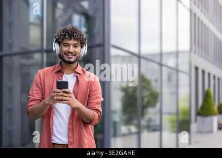 Porträt eines jungen Indianers mit Kopfhörern, der durch die Stadt geht, ein Telefon hält und Musik hört. Stockfoto