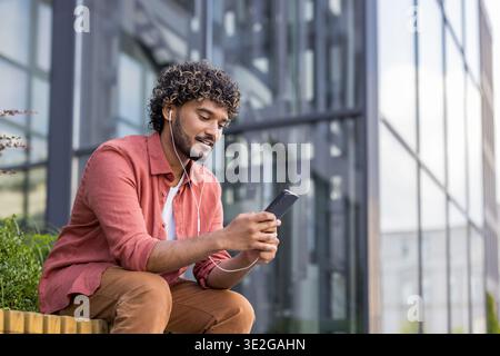 Ein junger Indianer in lässiger Kleidung sitzt mit Kopfhörern auf einer Bank in der Nähe eines Gebäudes und benutzt sein Handy mit einem Lächeln. Stockfoto