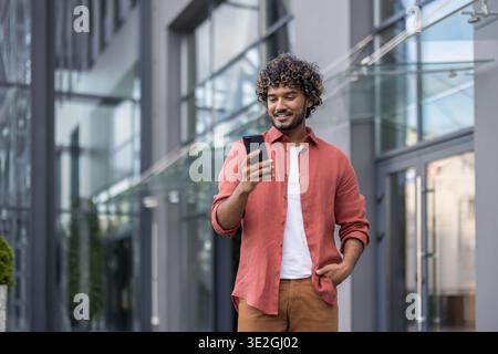 Junge lächelnde muslimische männliche Studentin, die auf dem Campus läuft, Hand in der Tasche hält und Handy benutzt. Stockfoto