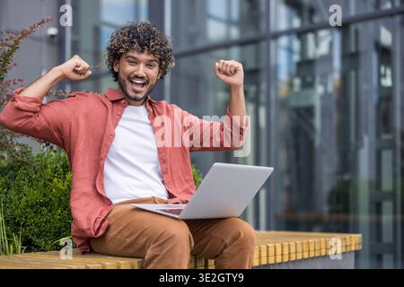 Porträt eines jungen Indianers, der draußen auf einer Bank sitzt, einen Laptop auf seinem Schoß hält und sich mit erhobenen Armen über den Erfolg freut. Stockfoto