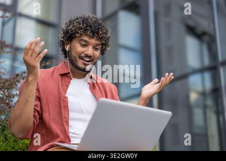 Nahaufnahme eines lächelnden jungen Indianers mit Kopfhörern, der draußen sitzt und auf einem Laptop spricht, den er auf seinem Schoß hält. Stockfoto