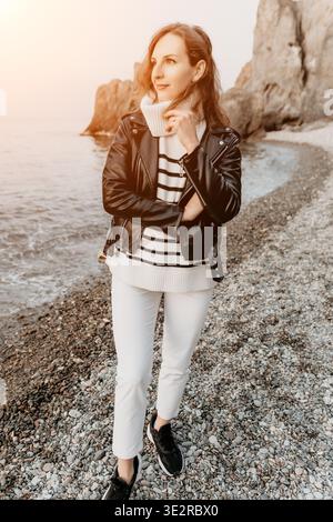 Frau Strandmode steht auf felsigen Kieseln in Lederjacke und gestreiftem Pullover und blickt auf das Meer bei Sonnenuntergang Stockfoto