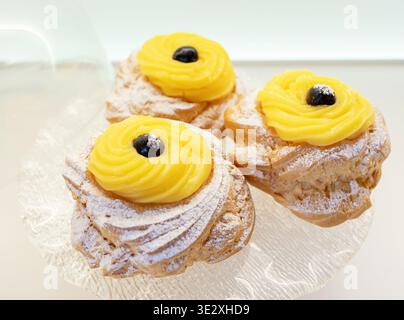 Zeppole di San Giuseppe mit traditionellem Choux-Gebäck, gekrönt mit lebendiger Gebäckcreme und einer wilden schwarzen Kirsche, die mit Puderzucker auf einer Glasplatte bestäubt wird Stockfoto