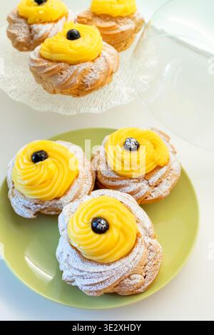 Traditionelles Zeppole di San Giuseppe Dessertgebäck auf einem Teller und einem Glasständer, garniert mit gelber Vanillecreme und dunkler Wildkirsche, mit Puderzucker bestäubt Stockfoto