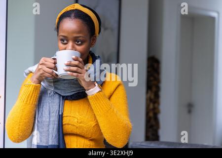 Afroamerikanerin mit weißem Becher, Senfpullover und Smartwatch zu Hause. Kontemplativ, gemütlich, gelb, Strick, Schal, Sonnenlicht, Wohnzimmer || Model veröffentlicht Stockfoto