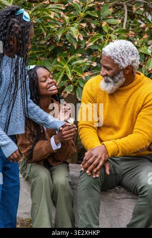 Afroamerikanischer Familiengroßvater mit Teenager, Kind, das auf Blockpullover sitzt und Gänseblümchen im Garten hält. Senior, Großvater, Enkel, Outdoor, Natur, Bank, grün || Modell veröffentlicht Stockfoto