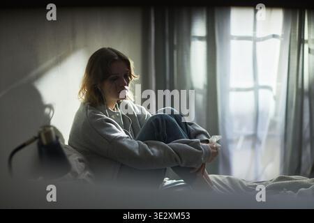 Kaukasische junge Erwachsene Frau sitzt auf dem Bett und umarmte Knie, hört Musik mit Ohrhörern, blickt mit traurigem Gesichtsausdruck nach unten, Sonnenlicht strömt durch Fenster im Hintergrund Stockfoto