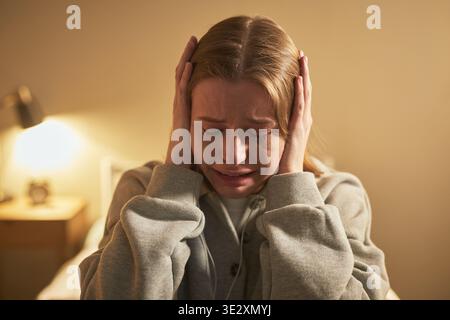 Porträt einer jungen erwachsenen kaukasischen Frau, die den Kopf in den Händen hält, mit geschlossenen Augen weint, intensive emotionale Not ausdrückt, drinnen mit neutralem Hintergrund sitzt Stockfoto