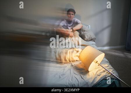 Junger Erwachsener kaukasischer Mann, der auf dem Bett sitzt und eine Decke mit angespanntem Ausdruck packt, die Lampe auf dem Boden umgeschlagen, die starke Emotionen und Ängste im Schlafzimmer vermittelt Stockfoto