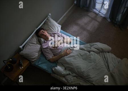 Kaukasischer junger Erwachsener, der im Bett liegt und ein Smartphone hält, mit nachdenklichem Ausdruck nach oben blickt, kontemplativ und emotional verloren wirkt, Schlafzimmerumgebung sichtbar Stockfoto