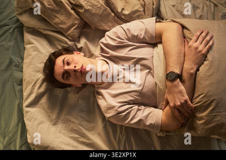 Porträt eines kaukasischen jungen erwachsenen Mannes, der auf dem Bett liegt und mit ernstem Ausdruck nach oben starrt, Arme über der Brust gekreuzt, Gefühle der Besinnung, Apathie oder Sorge vermittelt Stockfoto