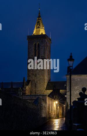 Ein Glockenturm aus leuchtendem Stein erhebt sich in der Abenddämmerung über einer schmalen, nassen Kopfsteinpflasterallee. Stockfoto